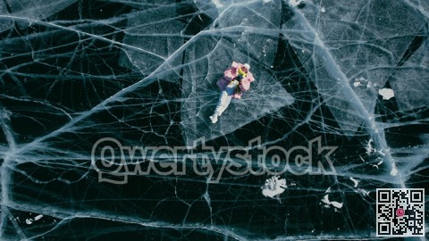 A tourist girl in bright clothes and sunglasses smiles happily, lies on the frozen blue ice of Lake Baikal and controls the drone up. An incredible pattern of c