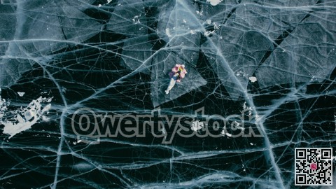 A tourist girl in bright clothes and sunglasses smiles happily, lies on the frozen blue ice of Lake Baikal and controls the drone up. An incredible pattern of c