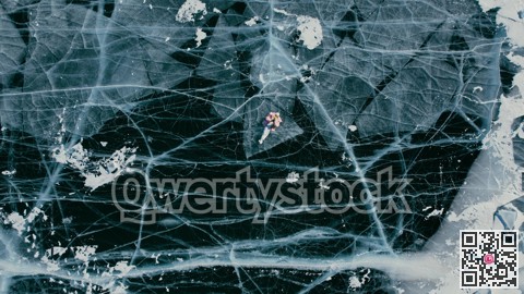 A tourist girl in bright clothes and sunglasses smiles happily, lies on the frozen blue ice of Lake Baikal and controls the drone up. An incredible pattern of c