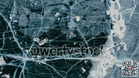 A tourist girl in bright clothes and sunglasses smiles happily, lies on the frozen blue ice of Lake Baikal and controls the drone up. An incredible pattern of c