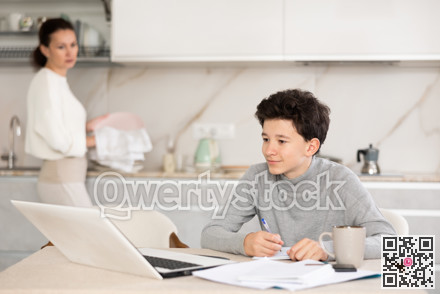 Teen son doing school homework while mom washes dishes in kitchen