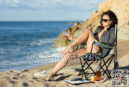 Naked woman in unbuttoned blazer sitting by ocean with whiskey