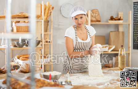 Skilled happy young woman baker in apron standing at work table, kneading dough for baking bread in bakery