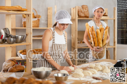 Skilled successful bakers in uniform working in bakery. Young girl working with pieces of dough and adult woman carries basket of baked goods