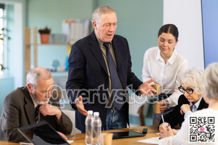 Elderly man standing and scolding sad senior colleagues at meeting table