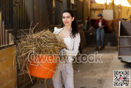 Armenian young female worker walks go through corridor of stable, carrying basket with straw