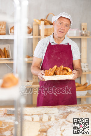 Smiling elderly baker in uniform stands behind counter in small private bakery offering croissants