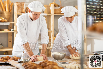 Two bakers, woman and young guy, knead raw dough on table in bakery kitchen