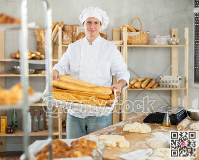 Positive young man baker in white uniform baking fresh baguettes in bakery
