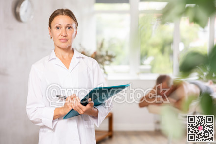 Positive middle-aged masseuse posing with folder in hand in the therapy room