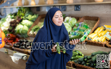 Delighted young Muslim woman purchaser choosing cucumber in grocery store
