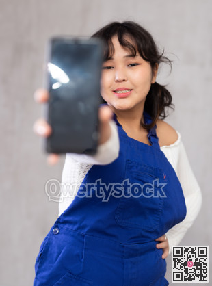 Positive girl in blue protective overalls showing modern smart phone with blank screen