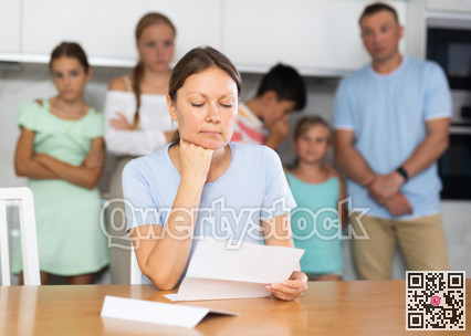 Upset mum is sitting in kitchen, reading letter. Blurred worried family in background