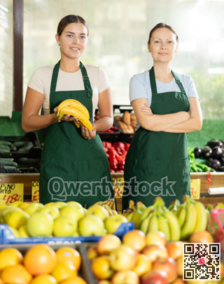 Friendly woman owner of fruit-vegetable shop standing in trading floor with girl assistant