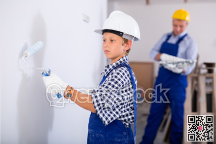 Boy in blue overalls paints the walls with paint roller