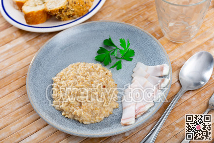 There is portion of boiled oatmeal porridge with pieces of salted bacon on plate.