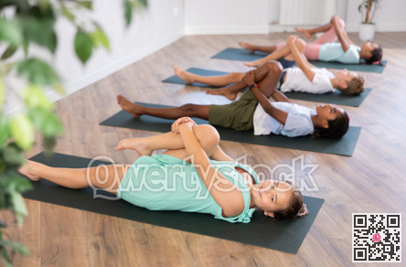 Children doing yoga with group in studio, lying on mat with knee pressed to chest with hands, performing hamstring and low back stretching Ardha Pawanmuktasana