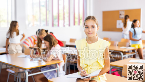 Portrait of schoolgirl with notebook in her hands who sits on desk in school class