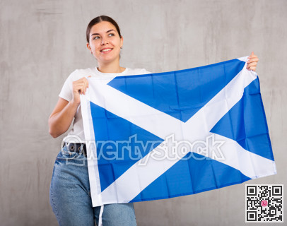 Happy young female student with flag of Scotland against gray wall background indoors