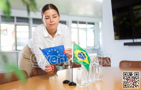 Positive young woman putting little flag of European Union on table next to the flag of Brazil