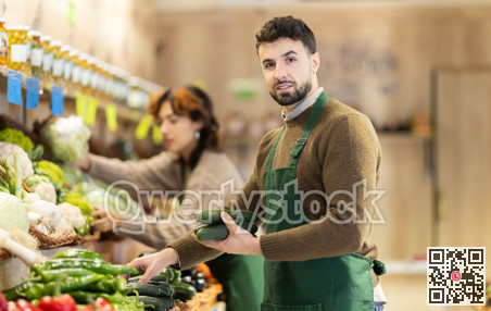 Young male seller putting cucumbers on counter in grocery market