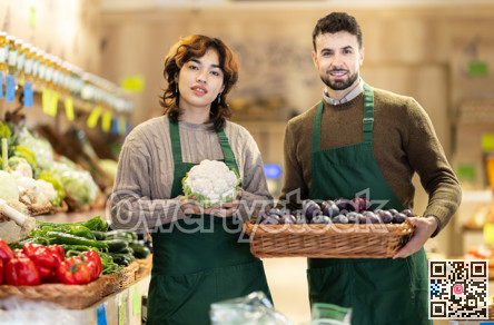 Young male and female sellers posing in grocery market holding cauliflower and plums