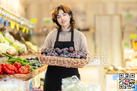 Young female seller holding basket of plums in grocery market