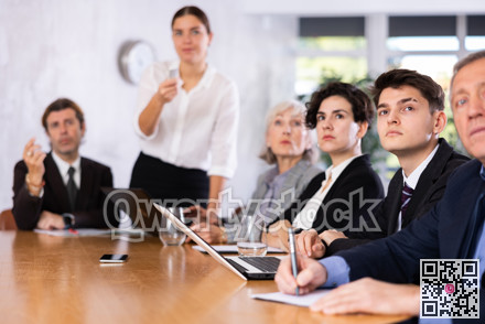 Office coworkers of different ages watching presentation while sitting at table with laptop and notes