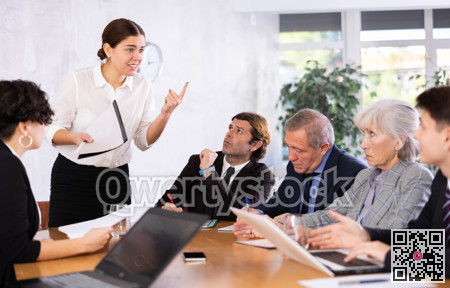 Clever young female making report while office workers of different ages using notebooks at table in office