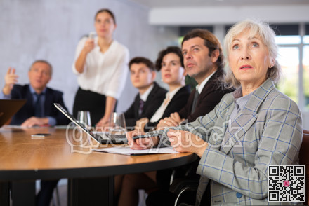 Office coworkers of different ages watching presentation while sitting at table with laptop and notes