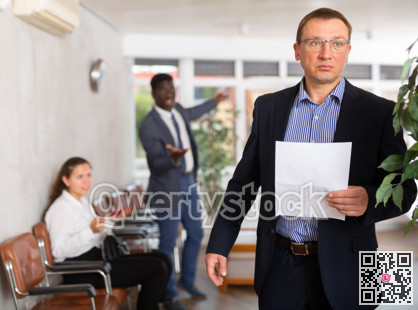 Upset middle-aged man standing in waiting room with his back to person who is shouting at him