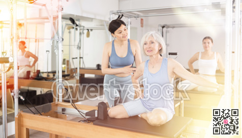Female trainer checking and helping senior woman exercising on pilates reformer machine in health center