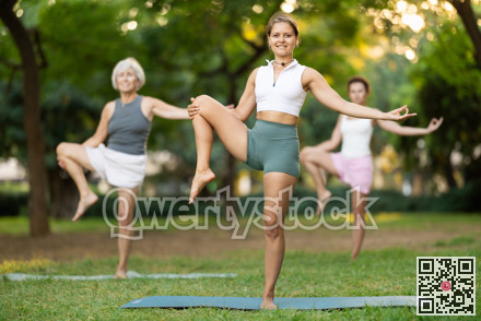 Girl in sportswear exercising with group people in park at daytime, standing in warrior one leg pose on fitness mat