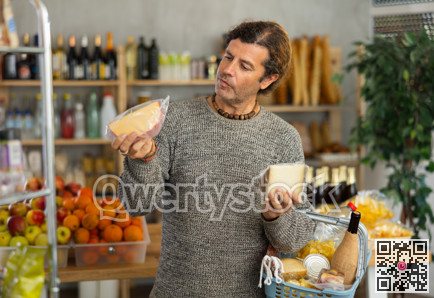 Attentive man with shopping basket trying to make choice holding two sorts of cheese in grocery store