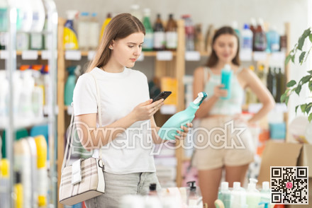 Young female shopper scans QR code on label of spray glass cleaner for windows using her smartphone, while in background shoppers browse products