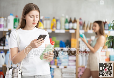Young female shopper scans QR code on label of dishwashing using her smartphone, while in background shoppers browse products