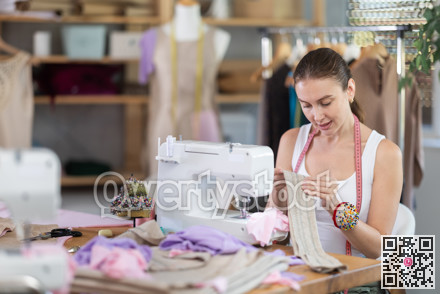 Glad young dressmaker viewing dress detail sitting at sewing-machine