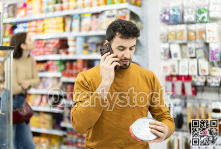 In store, man examines tin can product and talks on phone, advises on purchase.