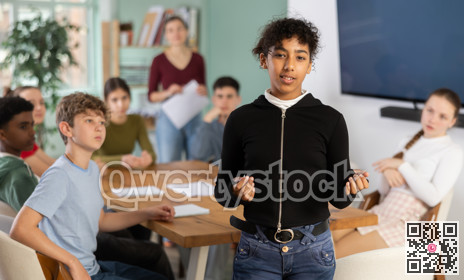 Glad teenage girl standing with her back to classmates sitting at table