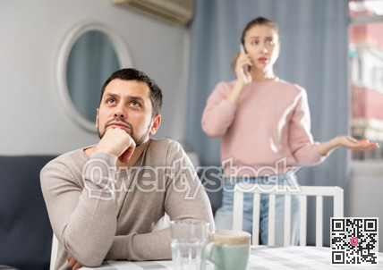 Sad man sitting at table at home, wife talking on phone