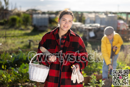Smiling young female farmer posing with basket and garden instruments during season of sowing on sunny day of autumn