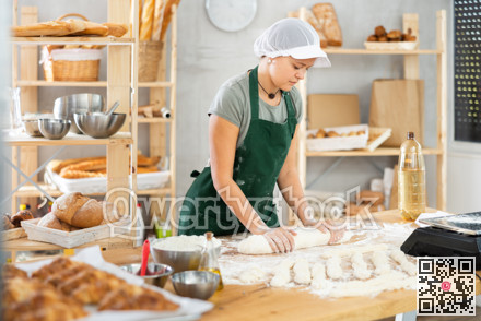 Young baker girl is standing near work table in kitchen, working with dough