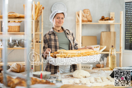 Bakery female seller stands proudly in front of fresh bread holding basket of freshly baked baguettes in kitchen