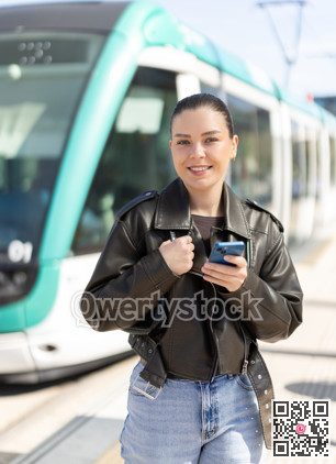 Young woman waiting for tram at stop with mobile phone