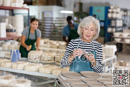 Portrait of elderly woman in apron cutting burrs off freshly made earthenware cups and plates with knife