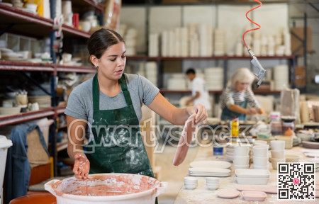 Young female worker in pottery factory lowers fired clay plates and cups into container with glaze