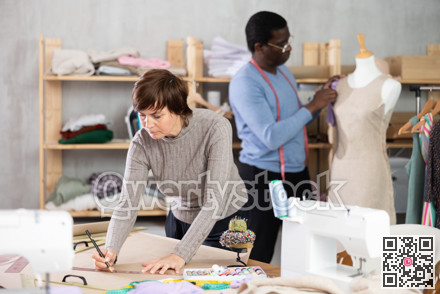 Female dressmaker sketching sewing pattern while colleague adjusting dress on mannequin