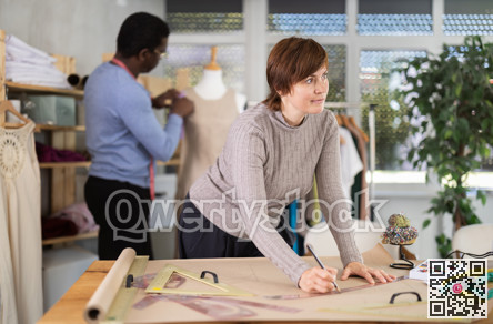 Woman draw dress pattern of product on paper. Companion work in background with mannequin