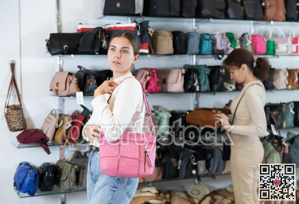 Girl stopped near accessories showcase, holds lady handbag in hands and examines product
