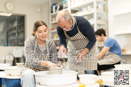 Eldery man helping young woman shape pottery on wheel